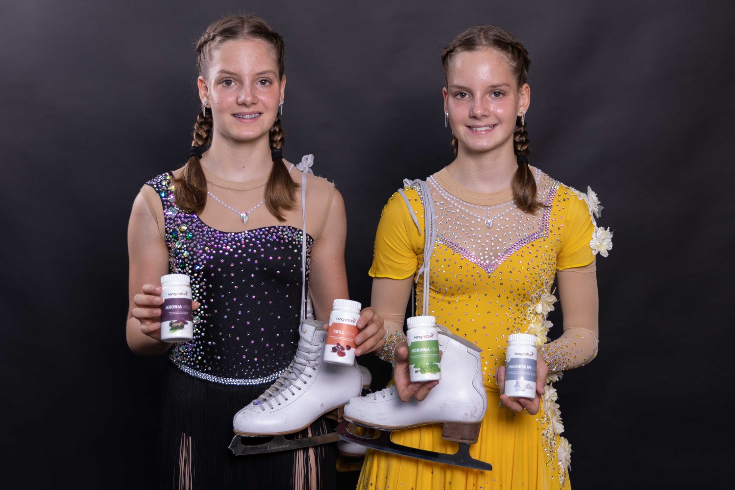 Zwei junge Eiskunstläuferinnen, Alessia Arnold und Selina Arnold, stehen in glitzernden schwarz-gelben Kleidern nebeneinander, halten weiße Schlittschuhe und verschiedene Nahrungsergänzungsmittelflaschen in den Händen und lächeln vor einem schwarzen Hintergrund.