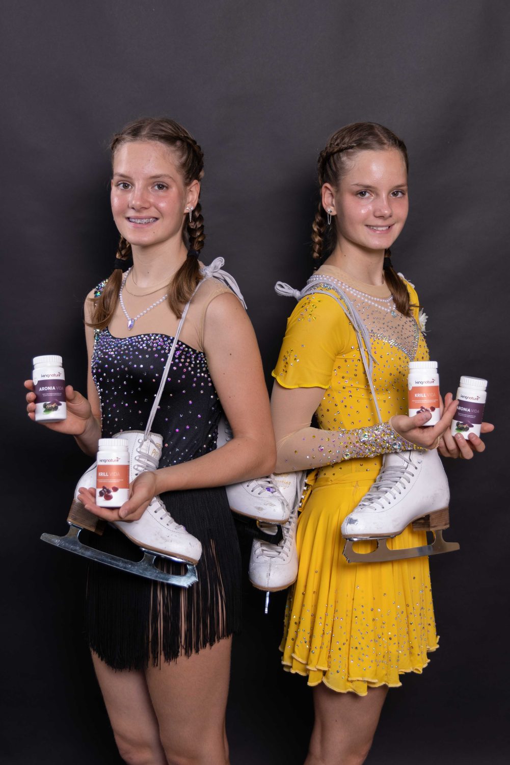 Zwei junge Eiskunstläuferinnen, Selina Arnold und Alessia Arnold, mit geflochtenen Haaren – die eine trägt ein schwarzes, die andere ein gelbes Kleid – stehen Rücken an Rücken und halten Flaschen mit Nahrungsergänzungsmitteln und Schlittschuhe vor einem dunklen Hintergrund. Beide lächeln in die Kamera.