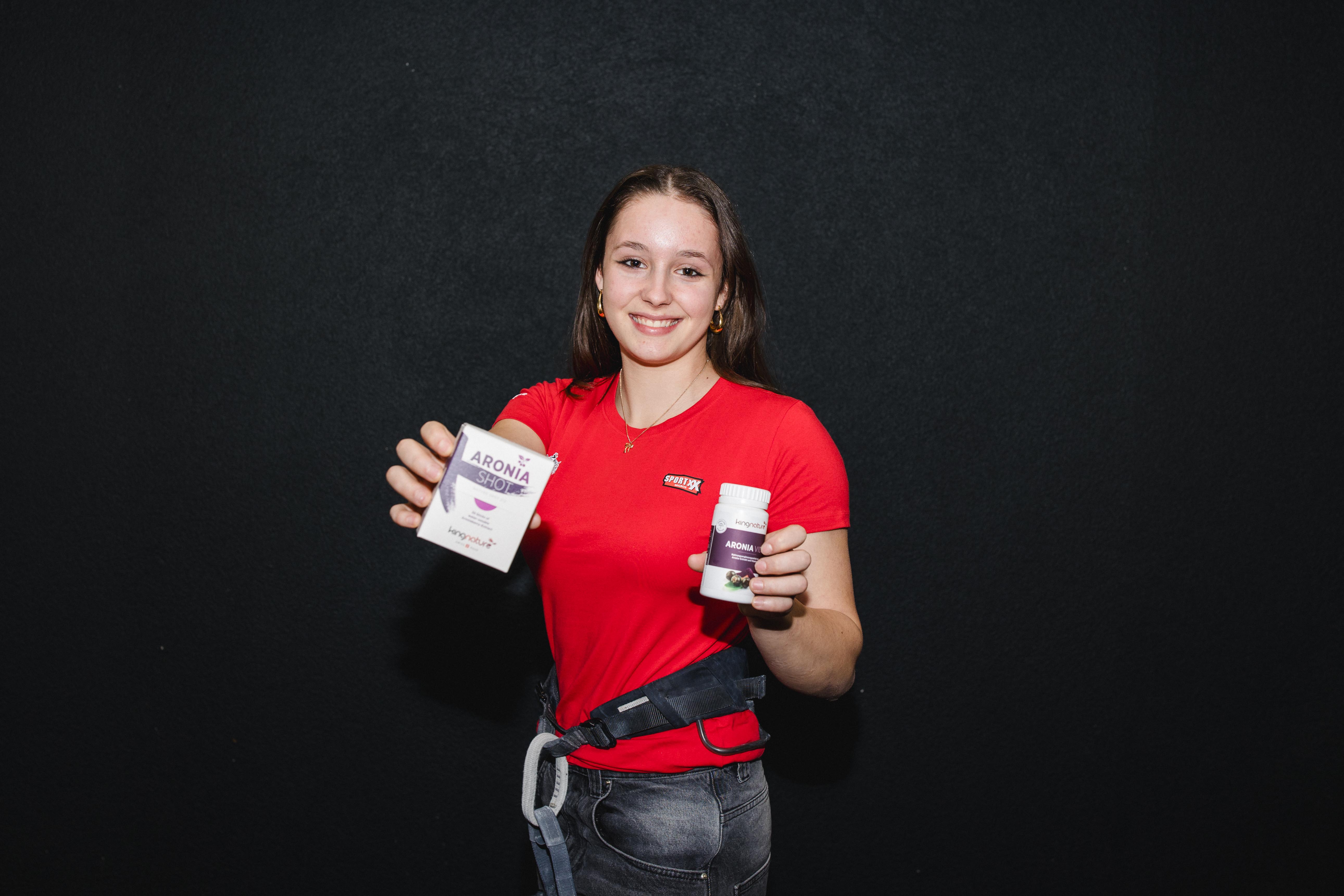Valentina von Gunten, eine junge Frau im roten Hemd, lächelt und hält vor einem dunklen Hintergrund eine Schachtel und eine Flasche Aronia-Nahrungsergänzungsmittel hoch.